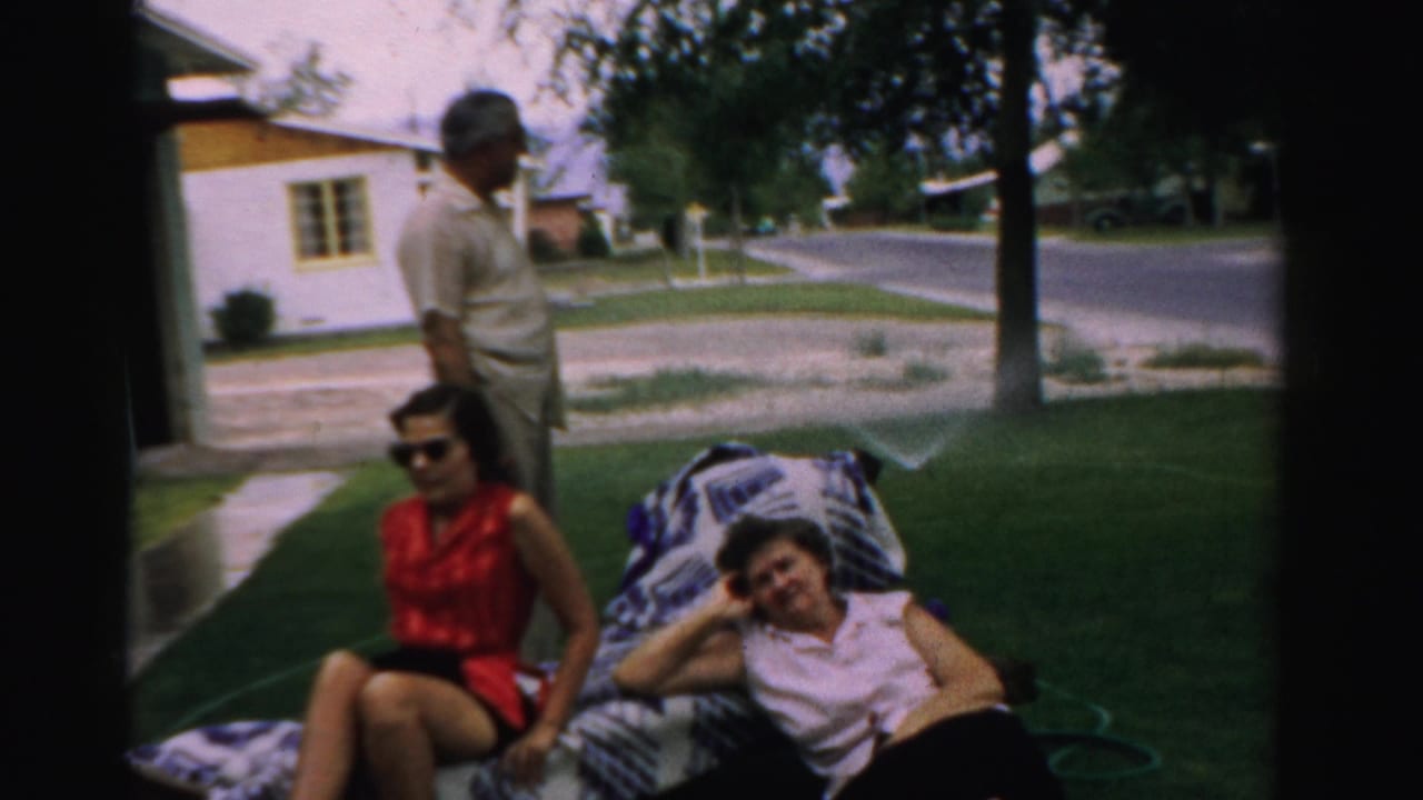 Man stands behind two women relaxing on a blanket in a suburban front yard.