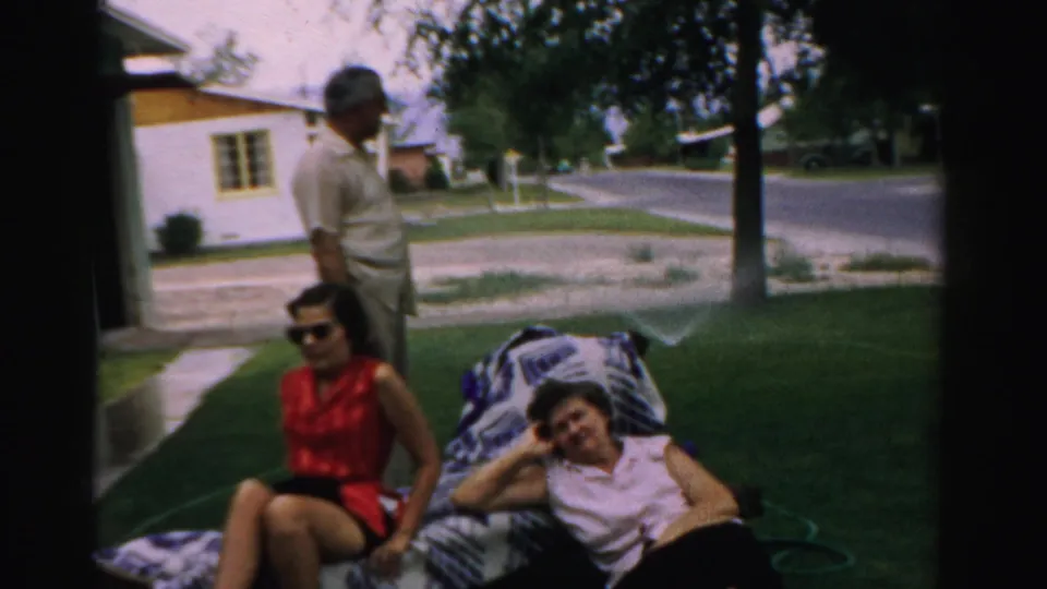 In 1957 Normal, Illinois, a man stands near two women on a patterned blanket in a grassy yard beside a quiet street.