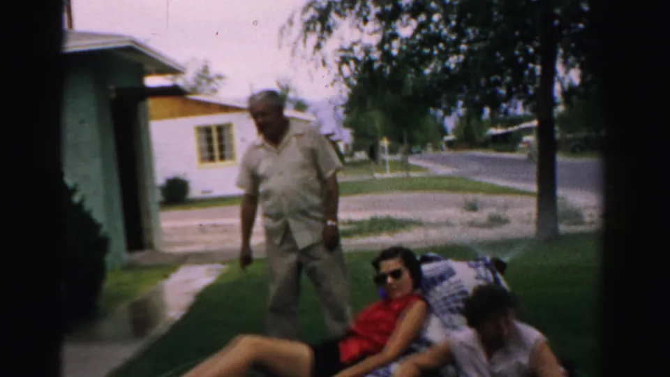 An older man in light clothing stands near two seated women on a blanket in a 1957 Normal, Illinois residential yard.