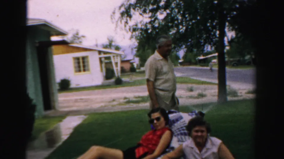 The man stands behind two women seated on a blanket in a shaded yard, with Normal, Illinois houses and street visible beyond.