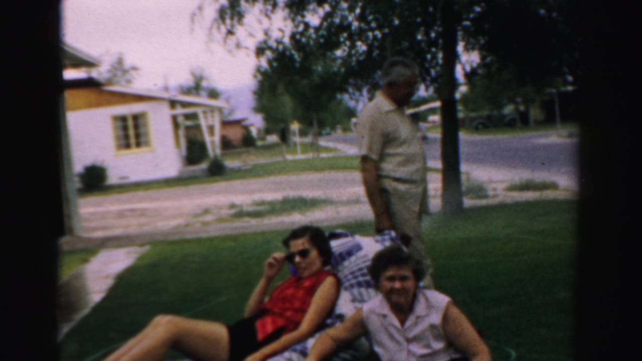 1957 Normal, Illinois home-movie frame showing two women relaxing on a lawn blanket as a man stands nearby in a suburban yard.