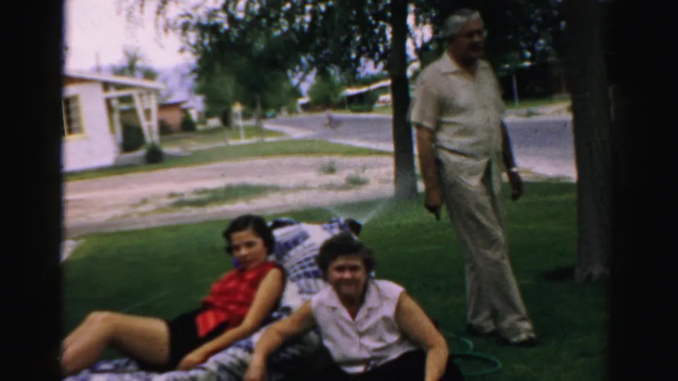 A home-movie frame from Normal, Illinois shows two women on a yard blanket and a man standing beside trees and a residential street.