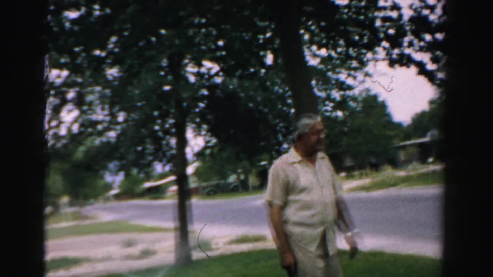 1957 home-movie frame showing a man standing in a shaded yard in Normal, Illinois, with trees, lawns, and a residential street behind him.