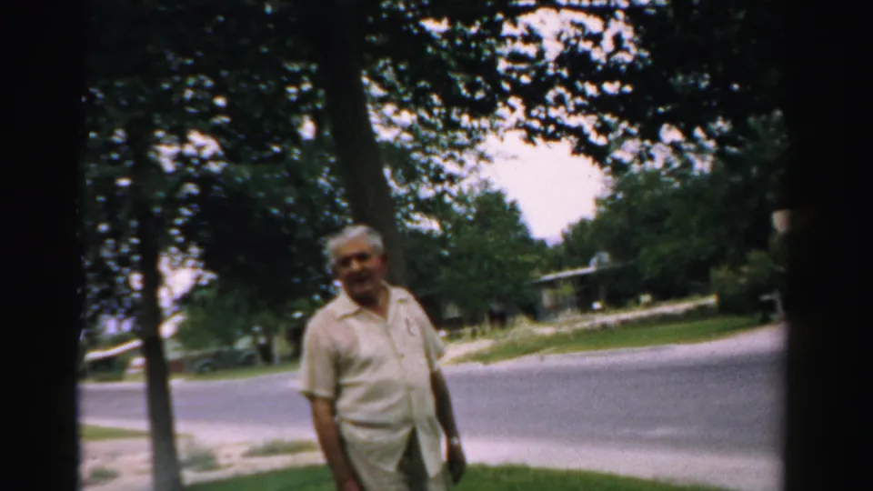 A late-1950s 8mm frame shows a man in a suburban Normal, Illinois yard, framed by trees and a quiet street.
