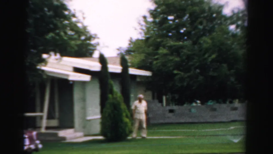 A wider 1957 Normal, Illinois yard view shows the woman near a low house, with lawn, trees, and shrubs around her.
