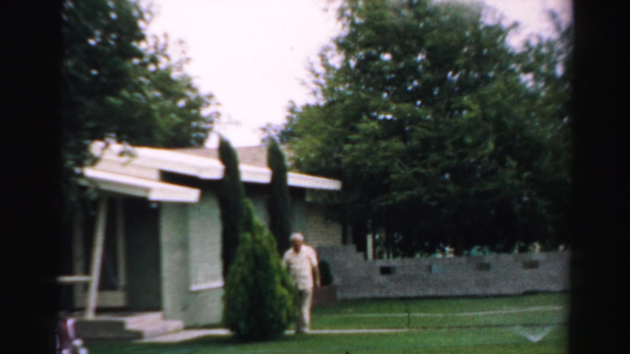 Elderly man standing beside a small suburban house and shrubs on a lawn.