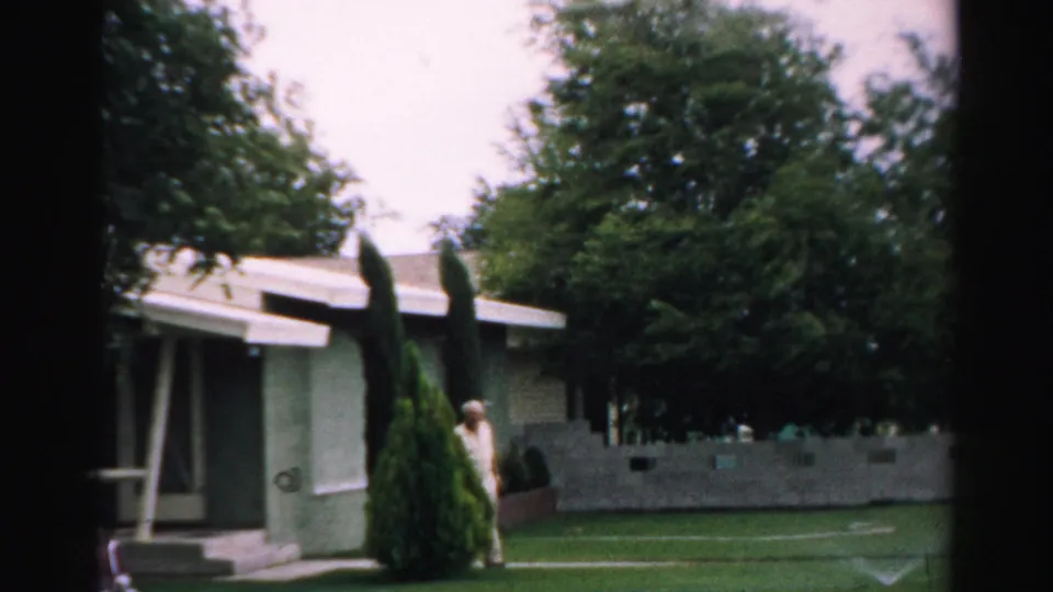 1957 Normal, Illinois exterior view of an older man partly near shrubs outside a low suburban house and yard.