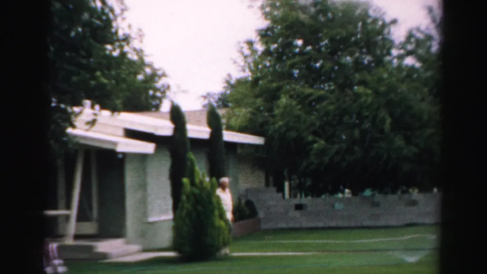 1957 Normal, Illinois frame with a modest house, lawn, evergreen shrubs, and an older man at the edge of the yard.