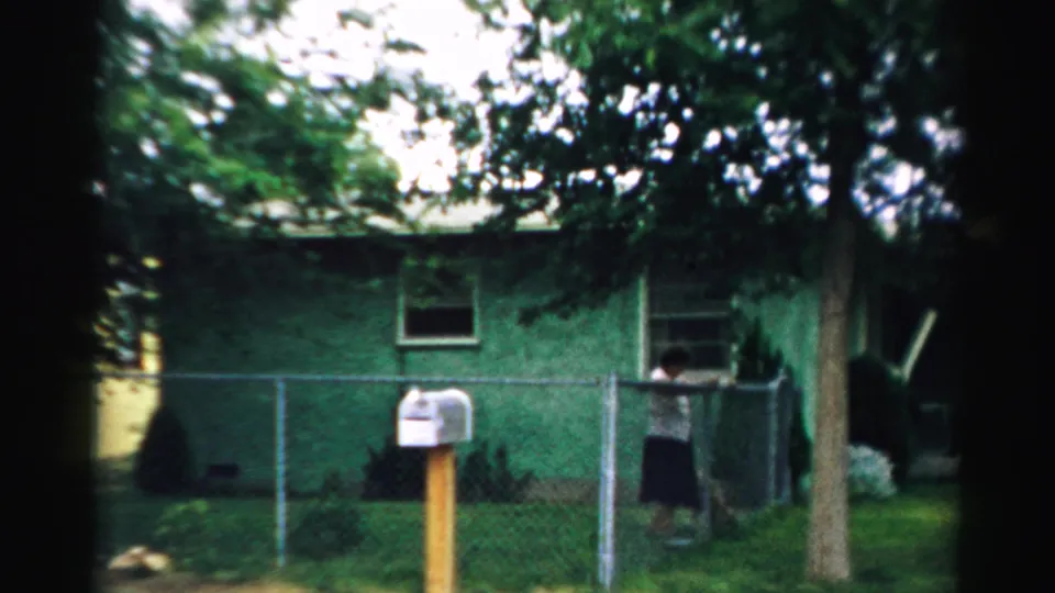 1957 Normal, Illinois backyard or side-yard view with a person near a chain-link fence, green house wall, mailbox, trees, and lawn.