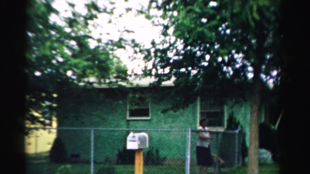 1957 Normal, Illinois 8mm frame showing a green residential structure, chain-link fence, white mailbox, trees, and a person by the gate.