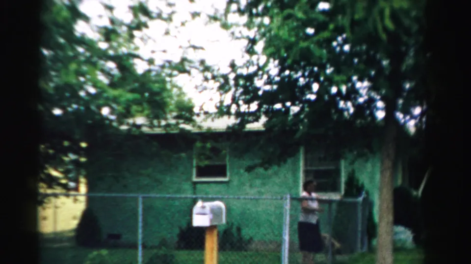 1957 Normal, Illinois 8mm frame showing a green residential structure, chain-link fence, white mailbox, trees, and a person by the gate.