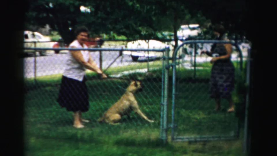 A woman stands with the seated dog in the fenced backyard, while another adult appears near the gate in the 1957 home-movie footage.