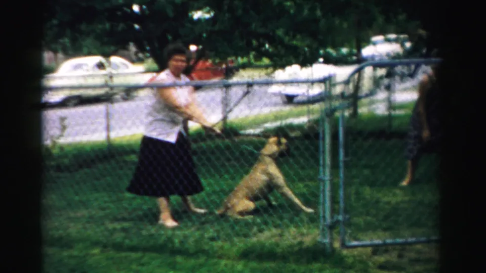 In 1957 Normal, Illinois home-movie footage, a woman in a white blouse stands with a light-colored dog in a grassy fenced yard.