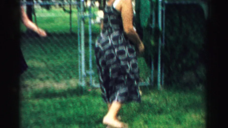 A barefoot woman in a patterned sleeveless dress crosses a grassy yard near chain-link fencing in the 1957 home-movie scene.