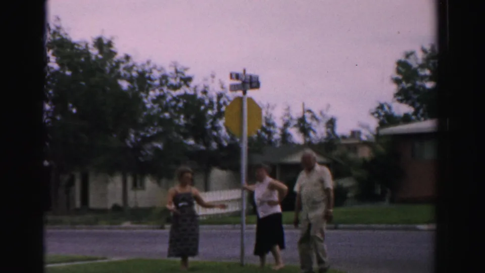Three adults are seen near a stop sign and curb in a 1957 Normal, Illinois suburban street scene.