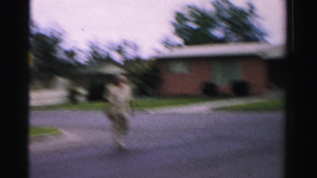 Soft 1957 home-movie frame from Normal, Illinois, showing a man crossing a residential street near brick houses and lawns.