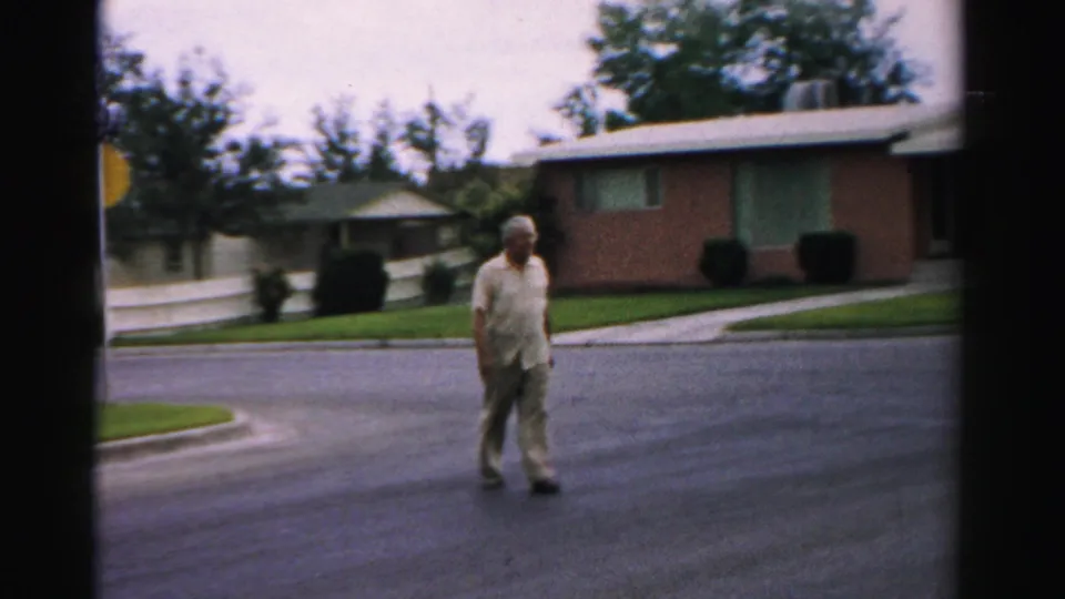 Older man in light clothing crosses a residential street in Normal, Illinois, with a brick ranch house and trimmed lawn behind him.