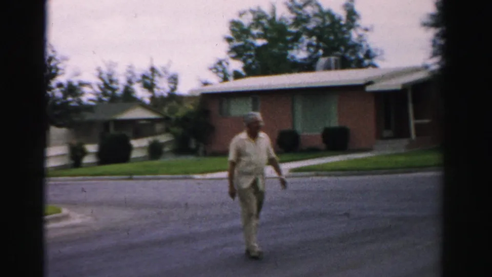 A man walks toward the camera on a quiet Normal, Illinois street, with lawns, shrubs, and brick houses in the background.