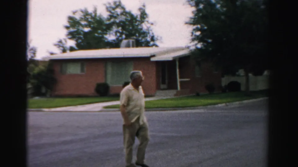 Man in light shirt and trousers passes closer to the camera on a residential street in 1957 Normal, Illinois.
