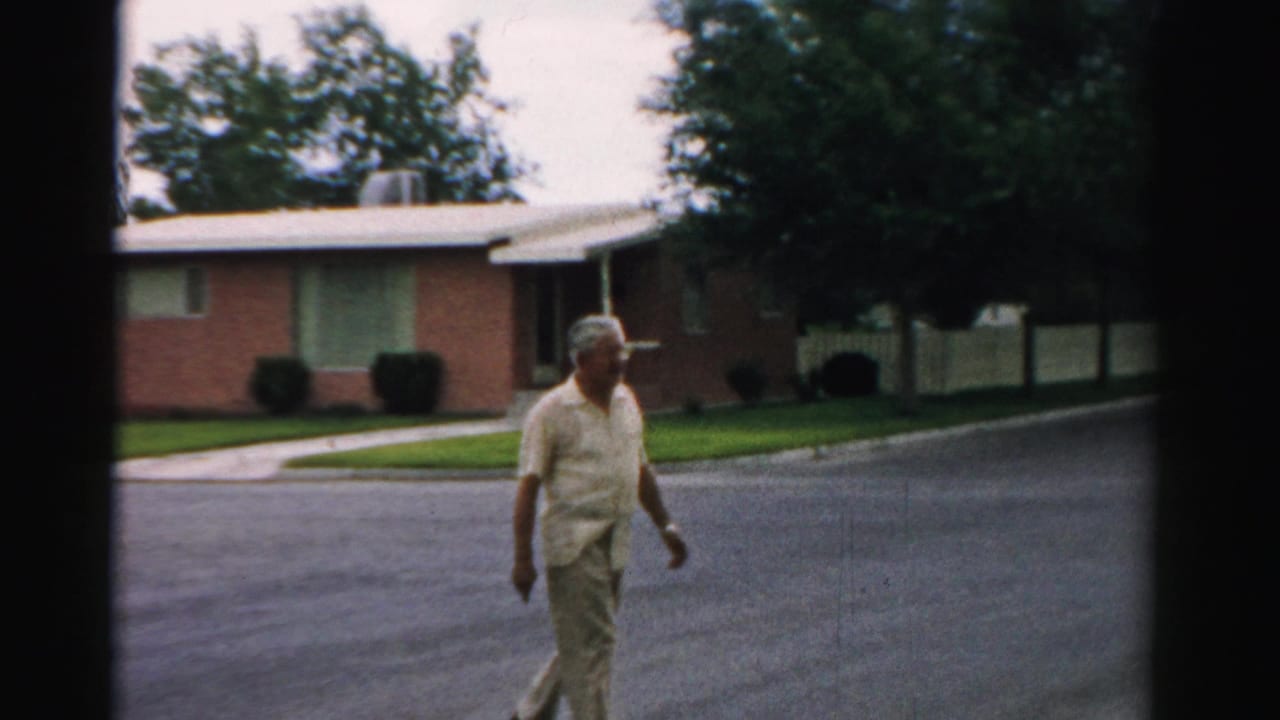 Older man walking across a suburban street near a brick ranch house and lawn.