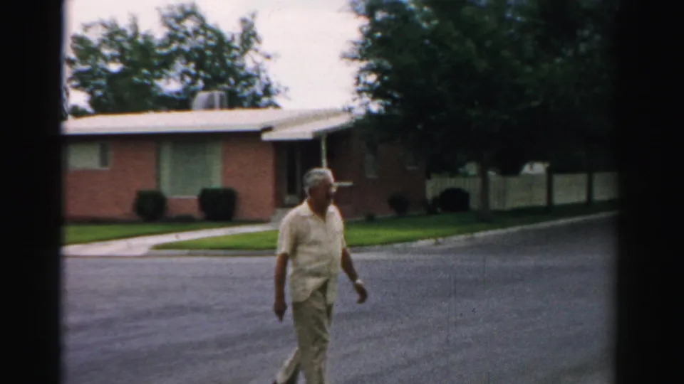 In Normal, Illinois, 1957, an older man in light clothing crosses a paved street beside a brick ranch home and trimmed lawn.