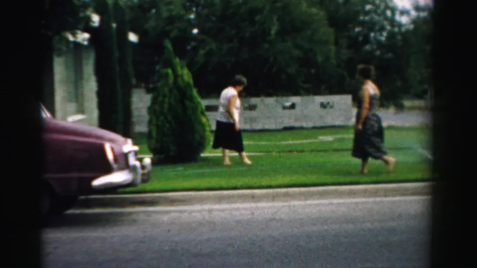 Two women walk on a suburban front lawn in Normal, Illinois, with a maroon 1950s sedan parked along the curb.