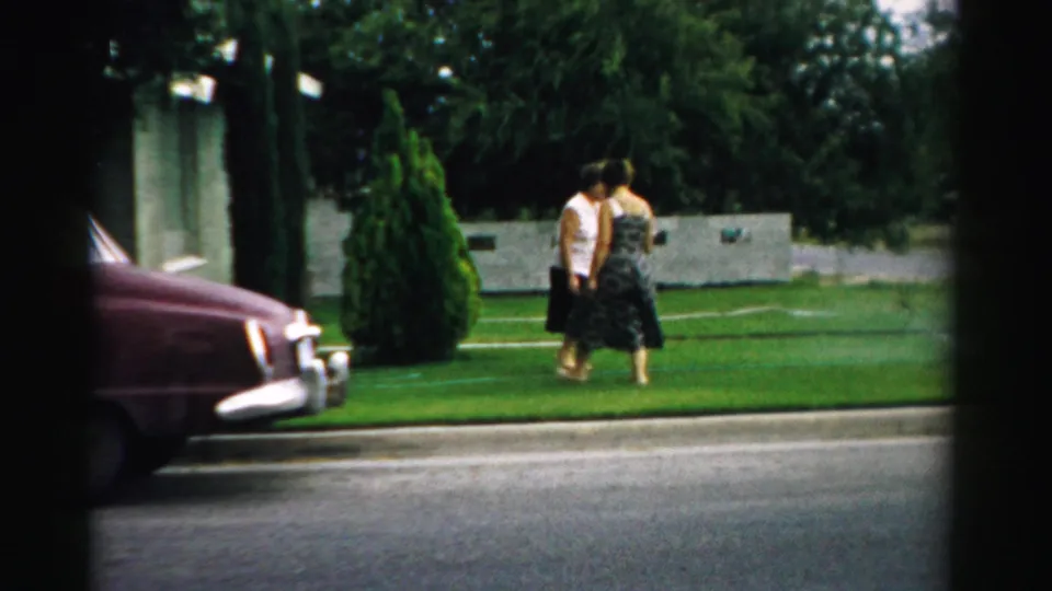 The women pass close together on a green front lawn in the 1957 Normal, Illinois home-movie scene, with the car nose at left.
