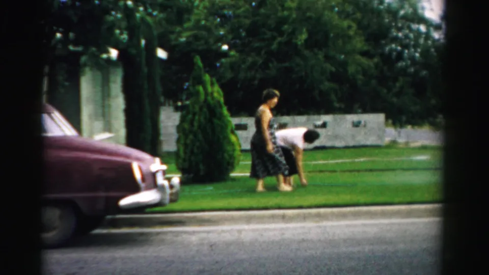 One woman continues across the Normal, Illinois front lawn while another bends toward the grass near the curb and parked sedan.