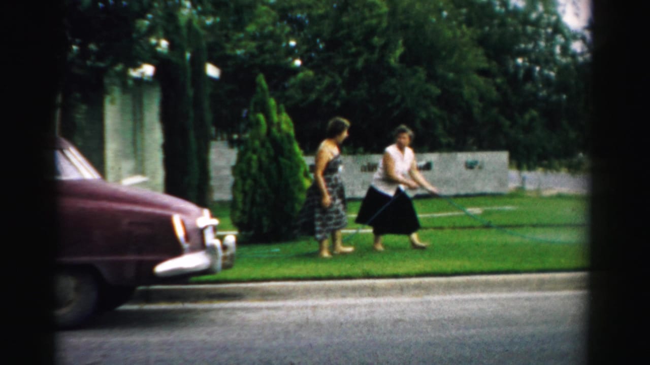 Two women stand on a green lawn near a garden hose, with a maroon car parked at the curb.