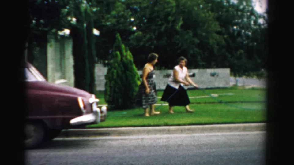 In Normal, Illinois in 1957, two women are on a suburban lawn beside a hose as a maroon car sits along the street.