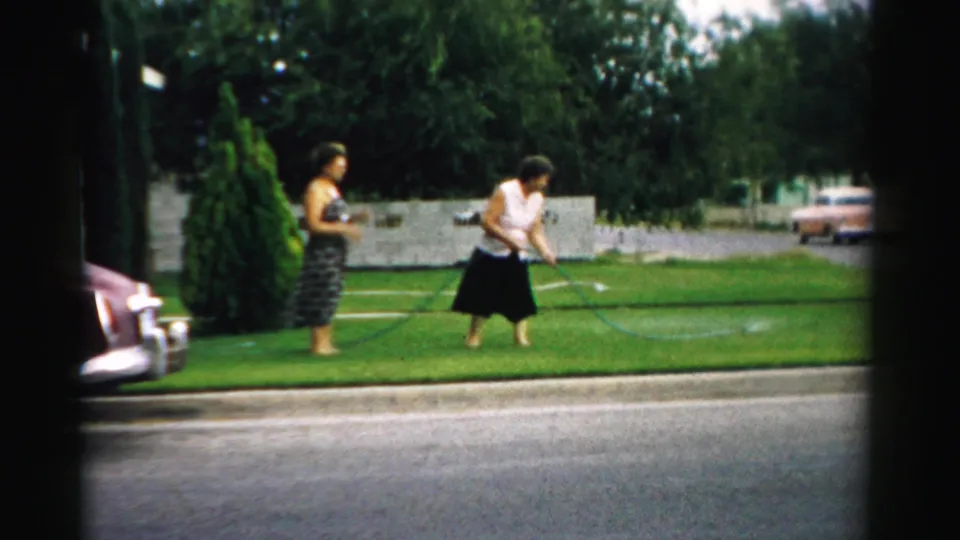 A 1957 Normal, Illinois home-movie frame shows one woman using a garden hose on a lawn while another watches nearby.