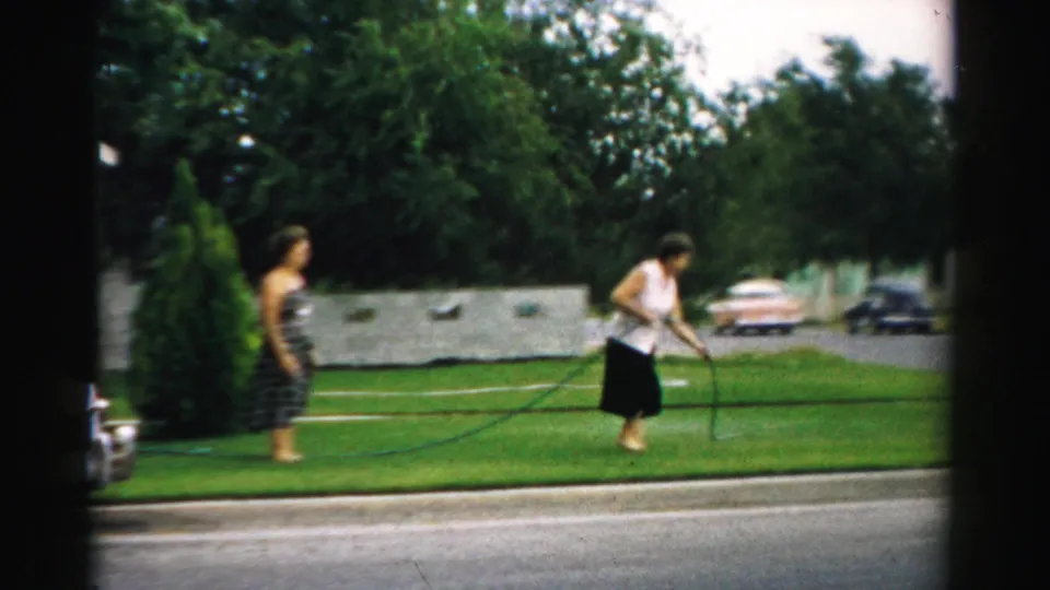In the residential Illinois yard, one woman bends slightly with the hose while another stands back near the edge of the lawn.