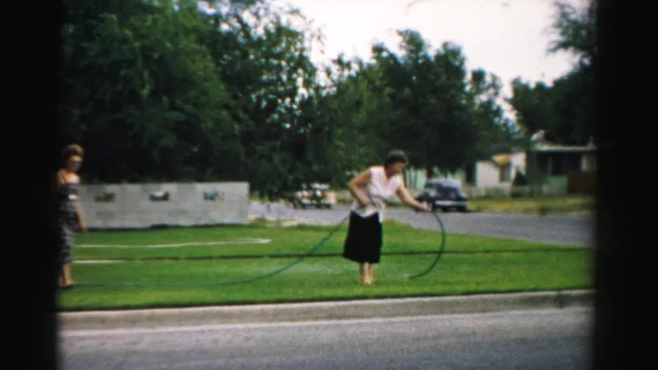 The 1957 home-movie frame shows a woman watering a suburban lawn in Normal, Illinois, with another woman partly framed at left.