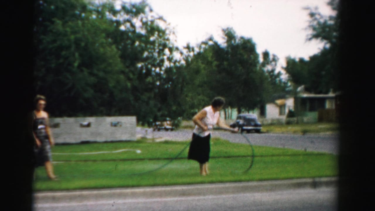 1957 Normal, Illinois home-movie frame showing two adult women on a green lawn, with one woman trying to manage a garden hose.