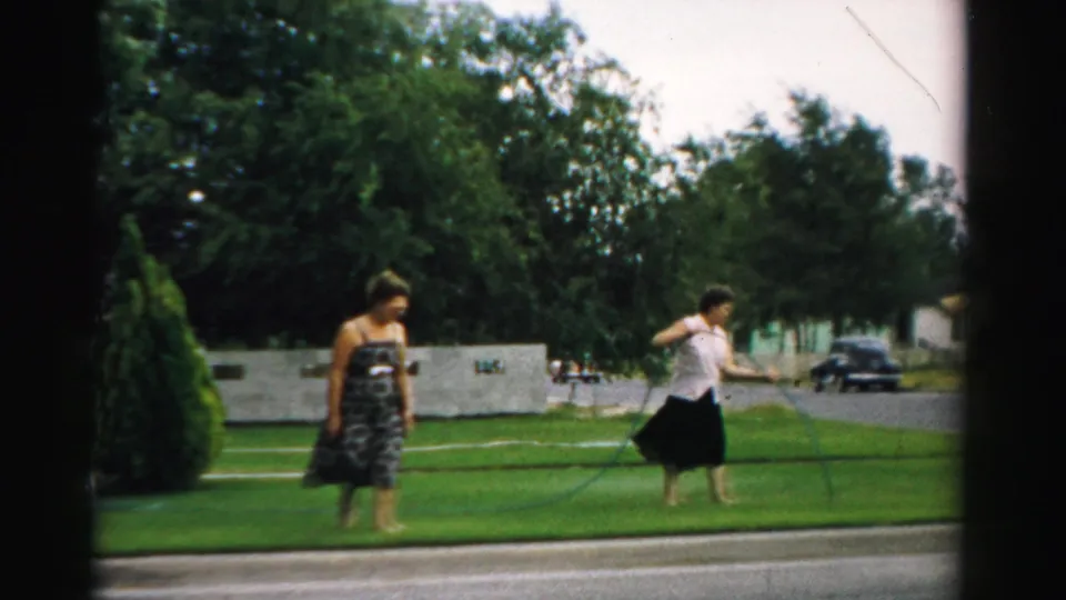 In a Normal, Illinois yard, a woman in a white blouse and dark skirt pulls a hose as another woman in a patterned dress crosses the lawn.