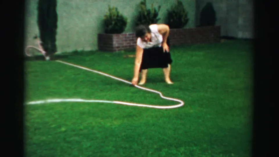 The woman bends low over a kinked garden hose on the lawn, with shrubs and a pale wall behind her in the residential yard.