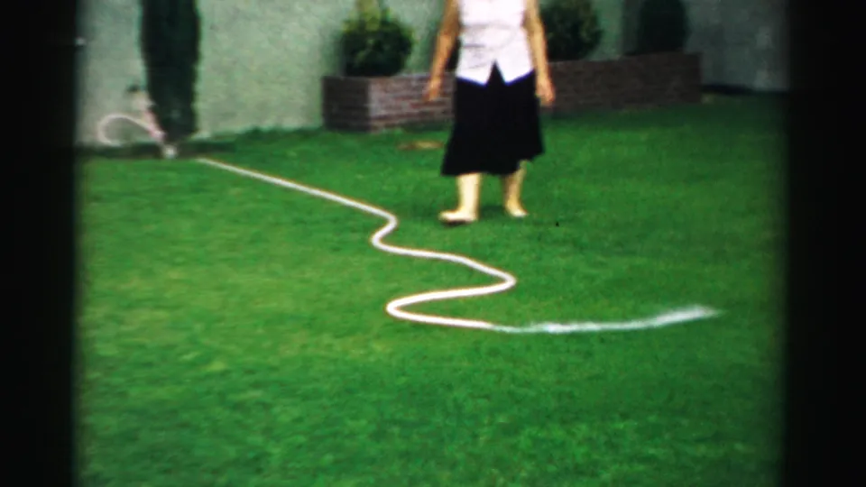 The woman stands barefoot or in light shoes near a looping garden hose, with a visible spray of water across the grass.