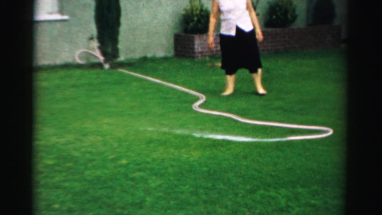 In a 1957 Normal, Illinois backyard scene, a barefoot woman in a white blouse and dark skirt watches a hose spray across the lawn.