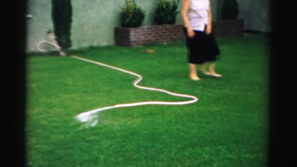 The hose sends water across the grass as the woman stands at the edge of frame in the 1957 backyard home-movie scene.