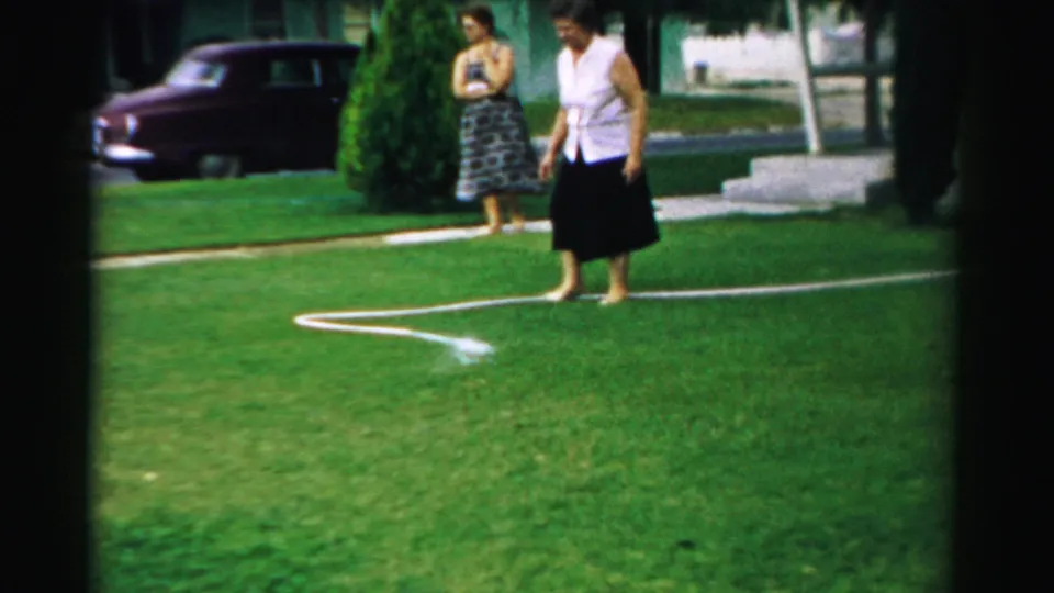 Two women stand in a suburban yard in Normal, Illinois, with a spraying hose on the grass and a mid-century car in the background.