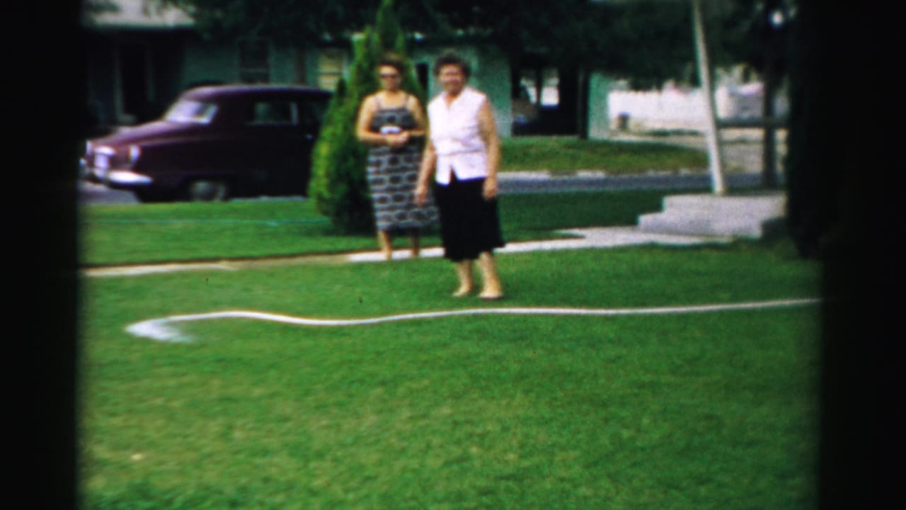 Two women stand on a green lawn near a garden hose with a maroon car parked behind them.