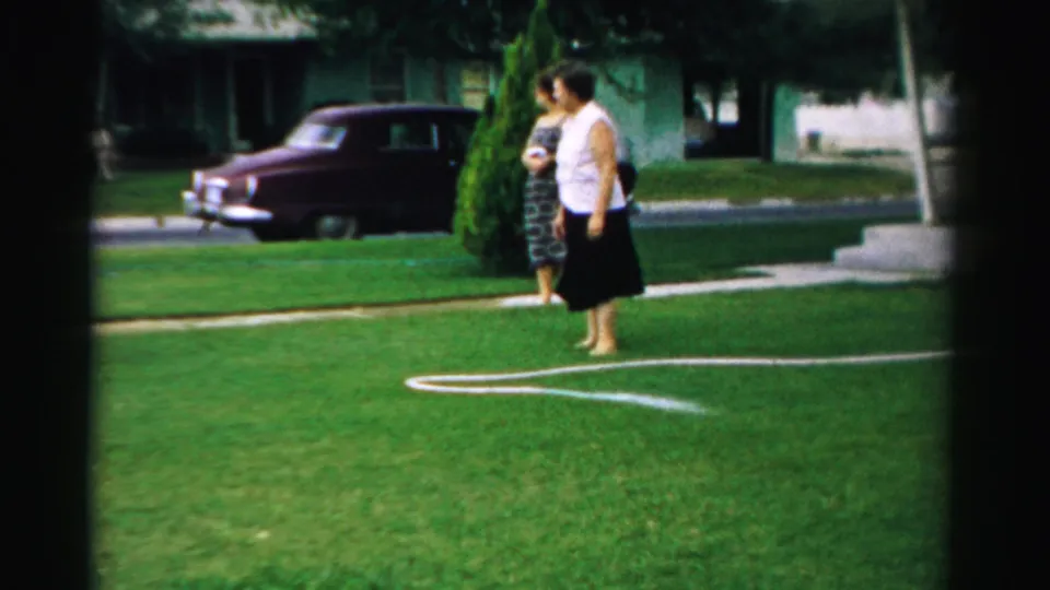 A barefoot woman in a white sleeveless top and dark skirt walks across a suburban lawn in Normal, Illinois, while another woman stands nearby.