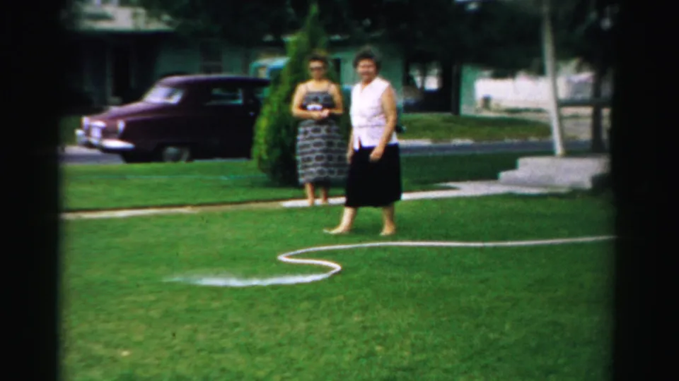 Two adult women are seen on a green suburban lawn in 1957 Normal, Illinois, with a white garden hose looping across the foreground.