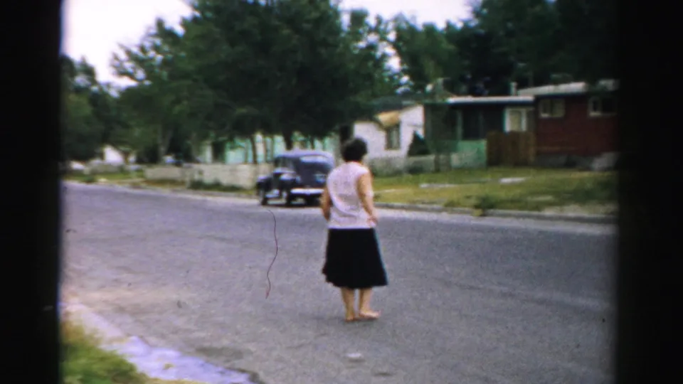 A barefoot woman in a white top and dark skirt stands in a Normal, Illinois residential street, with mid-century houses and a parked car behind her.