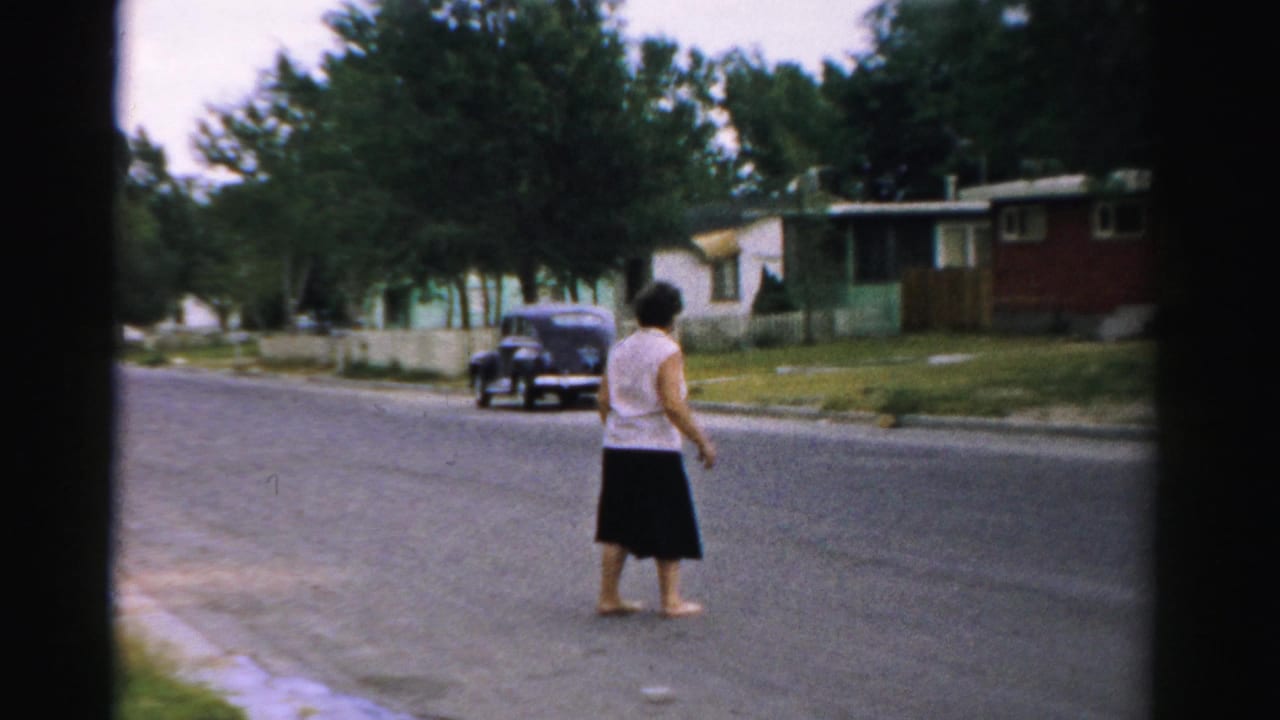 Woman walking barefoot across a quiet residential street with houses and a parked dark car nearby.