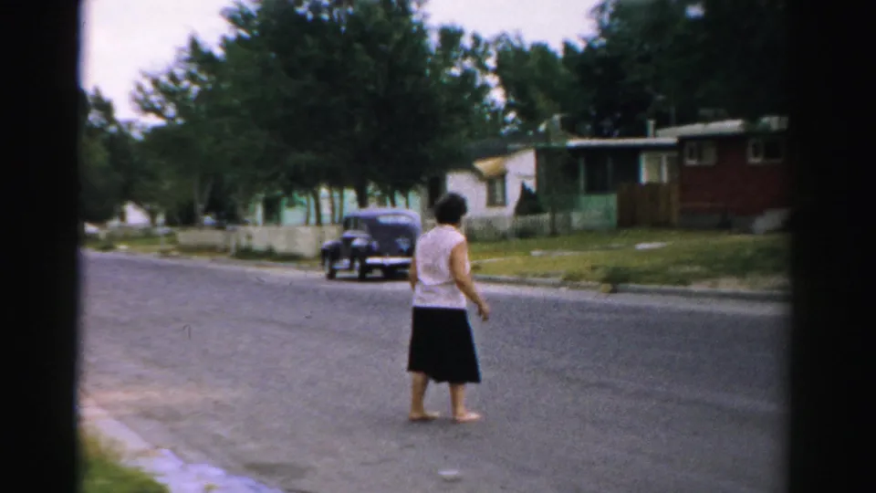 Adult woman in a white sleeveless top and dark skirt crosses a suburban street in Normal, Illinois, in 1957 home-movie footage.