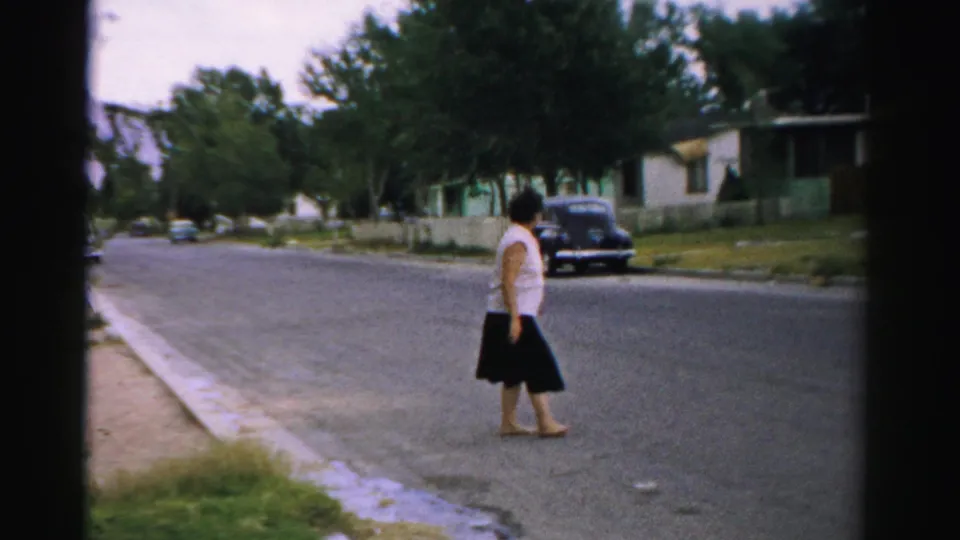 The woman pauses mid-street in a 1957 Normal, Illinois neighborhood, with low houses and a dark sedan along the curb.