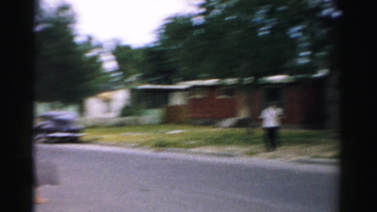 1957 Normal, Illinois home-movie frame showing a quiet residential street, trees, a distant adult, and a dark parked car.