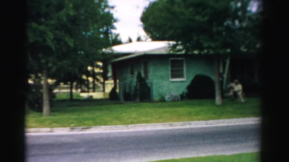 1957 Normal, Illinois home-movie frame of a green house, lawn, trees, and a distant seated person near the right side.