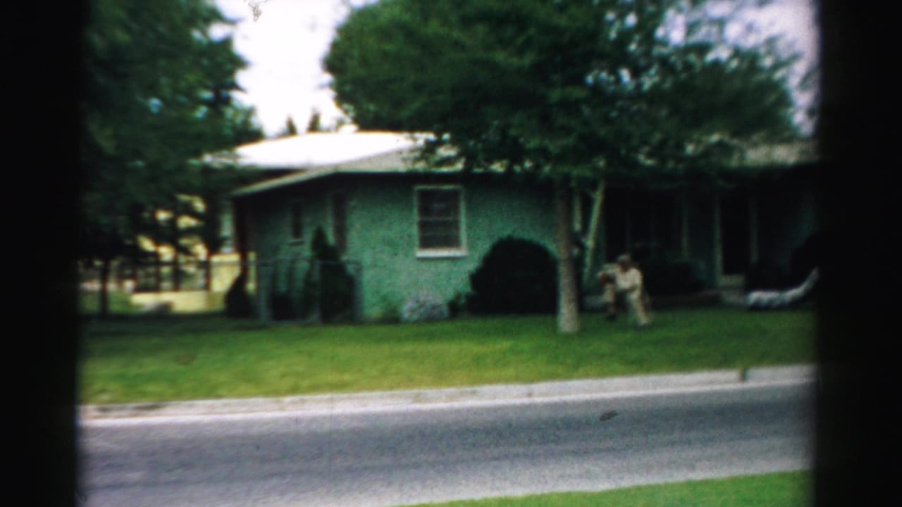 1957 home-movie frame from Normal, Illinois, showing a green single-story house, lawn, trees, and adults near the front entrance.